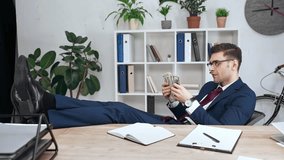 handsome business man counting and throwing dollar banknotes while sitting at workplace with legs on desk