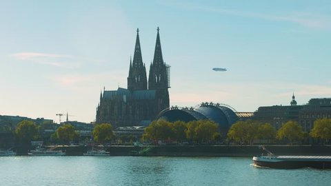 Cologne cathedral with flying Zeppelin and cargo ship on Rhine river in foreground, blue sky with clouds, green trees at riverside