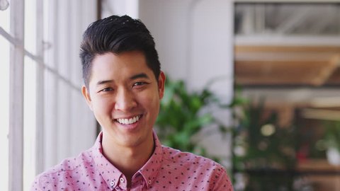 Millennial Asian male creative standing in an office turning and smiling to camera, close up