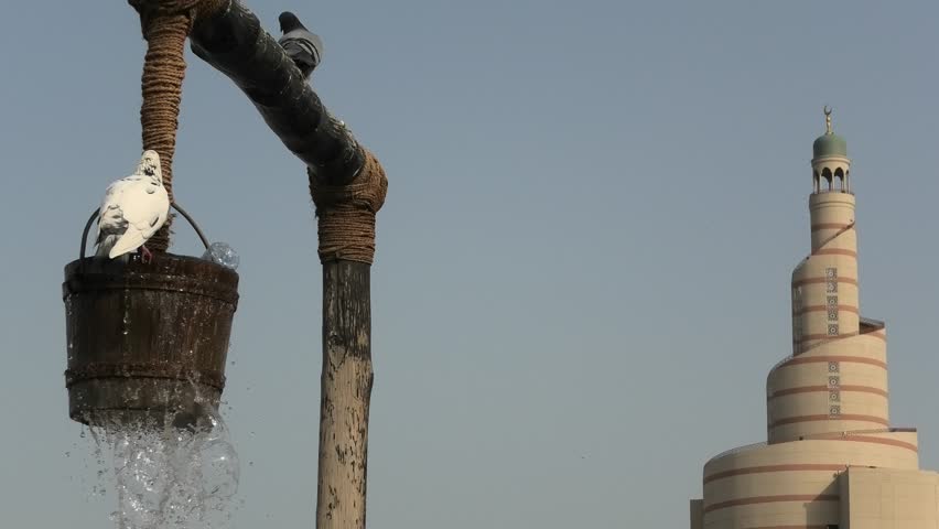 Pigeon drinks water at old well fountain, iconic landmark in the middle of Souq Waqif in Doha city center, Qatar. Middle East, Arabian Peninsula. Sunny blue sky. Doha Mosque on blurred background.