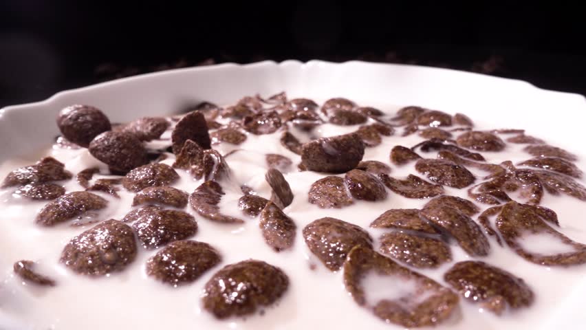 Brown chocolate corn flakes fall in a white plate with a pattern of flowers on the edge and are filled with milk. Closeup. 