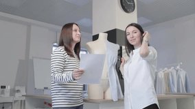 two girls of the fashion designer discuss something looking at a sheet of paper in sewing salon, a sample, the sketch of a photo of model in hands, against there is a dummy with a sample of clothes