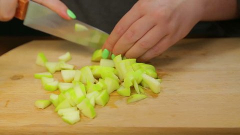 A woman cuts a green apple with a knife on a wooden board into small cubes. Time laps.