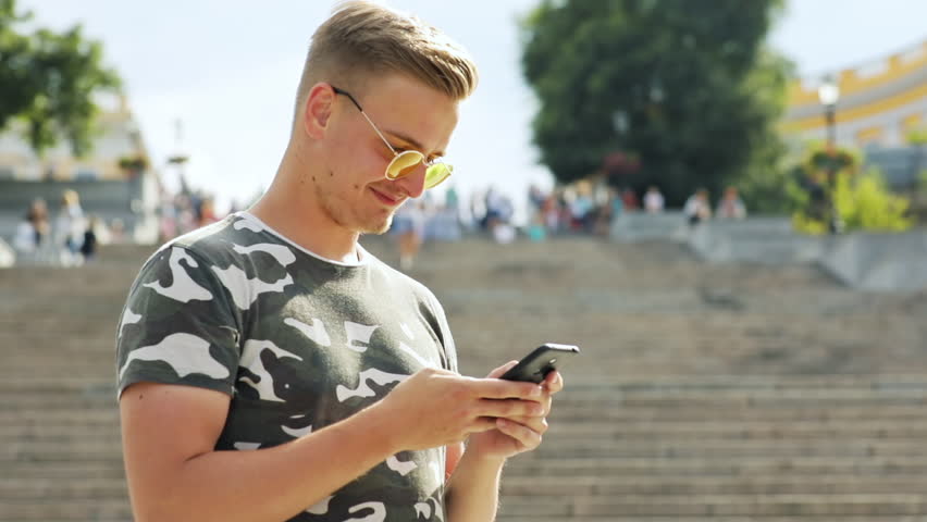 Handsome guy wearing sunglasses looking at smartphone standing against huge stairway in the city center in sunny weather.