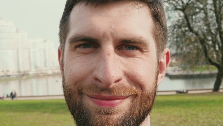 bearded young man portrait who smiling and then looking around on summer city background slow motion close up video in 4K