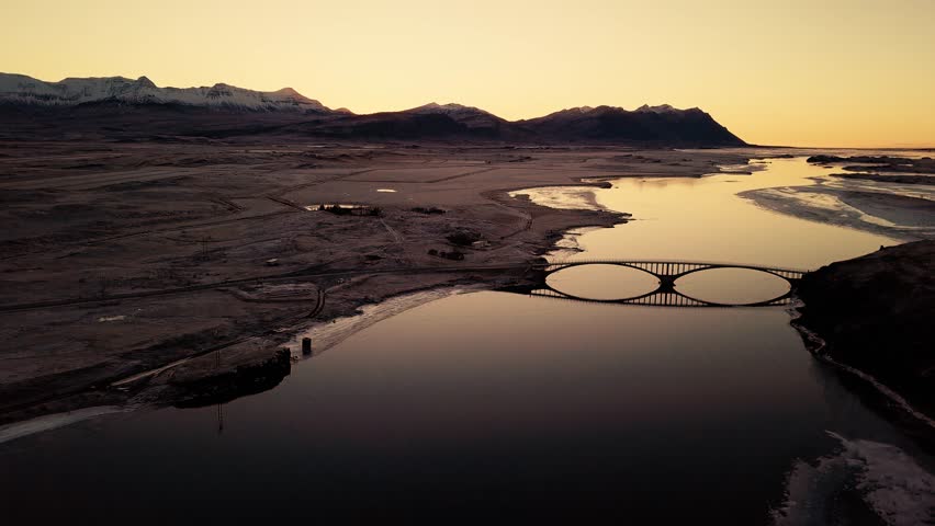 Drone flying towards a bridge being reflected in the river during sunset.