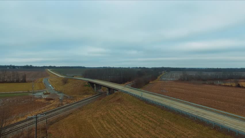 Cars, trucks and a bus traveling across an overpass running over railroad tracks next to a pond on a rural Illinois highway.
