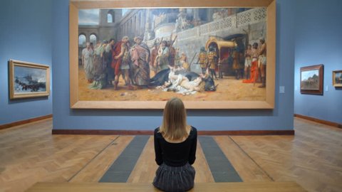 WARSAW, POLAND - MARCH 28, 2017: National Museum in Warsaw. Young Woman in a Dress with Straight blond Hair is Sitting on a Bench in the Museum or Art Gallery and Looking at a Huge Picture.