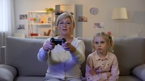 Excited granny playing video game ignoring sad grandchild sitting sofa addiction