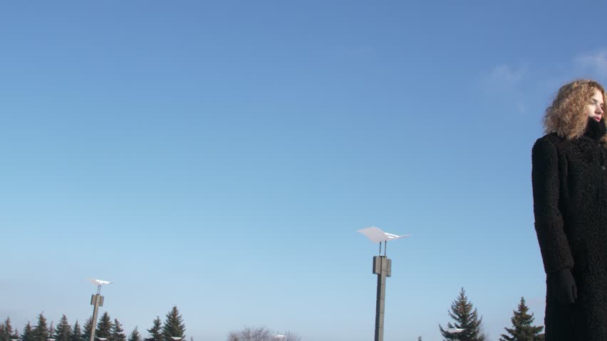 young woman in black winter coat standing in front of blue sky tracking camera movement