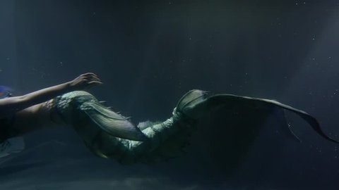 Side view shot of a model dressed as mermaid swimming by, swinging her big tailfin, dark underwater.
