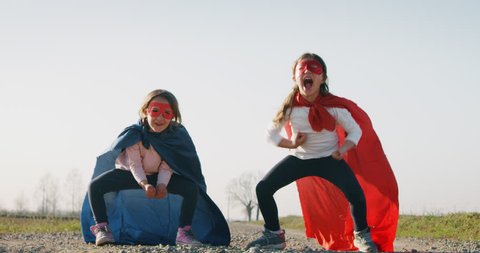 Authentic shot of a two funny little girls sisters dressed as superheroes in red and blue dancing and having fun on a sunny day.