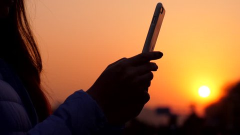 Woman texting using smartphone, silhouetted shot of mobile device in hands against vivid sunset. City street seen blurred on background, people stoll at sidewalk