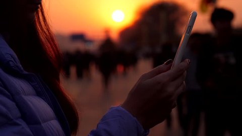 Smartphone in hands of woman, blurred street on background, people silhouettes and vivid orange sun light at sunset time. Dramatic natural lighting, tourist lady using phone, standing at sidewalk