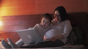 Happy Asian pregnant woman using laptop together with her daughter in bed, lovely Chinese little girl playing with her pregnant mother in the bedroom. happy family education lifestyle 4k clip
