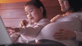 Happy Asian pregnant woman teaching her daughter using laptop in bed, lovely Chinese little girl learning playing with her pregnant mother in the bedroom. family education lifestyle slow motion clip