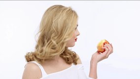 Young beautiful woman eating fruit and says yum. Happy adult girl tastes a red apple. VIdeo footage of a blond caucasian woman eats fruits over white background. 4k resolution.   