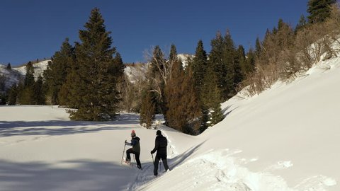Panning aerial view of couple snowshoeing through new snow on sunny day in the mountains.