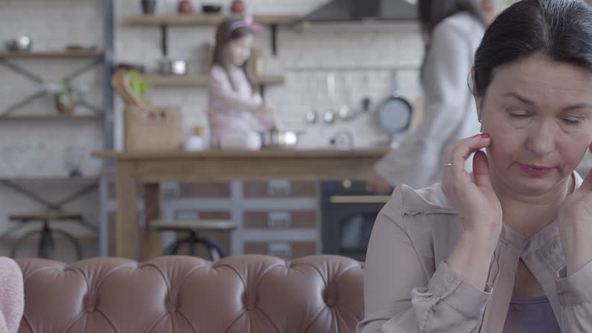 Sad senior woman sit on the sofa, crying and wipe her tears from the face. On the background her daugher and dranddaughter happy playing in the kitchen table.