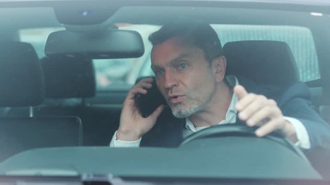 Close up view of handsome man in luxurious car impatiently beeping car horn while not moving in the traffic jam, then angrily talking on the phone and actively waving his arms. Stress, trouble