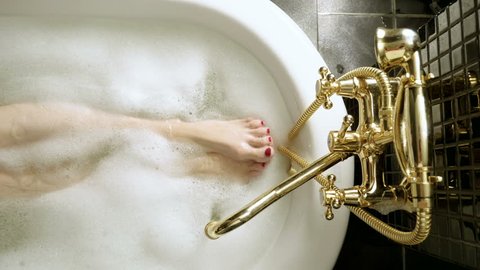 Top view of water flowing from golden faucet into the bathtub. The slim legs and feet with red pedicure of young woman taking a bubble bath. 4K