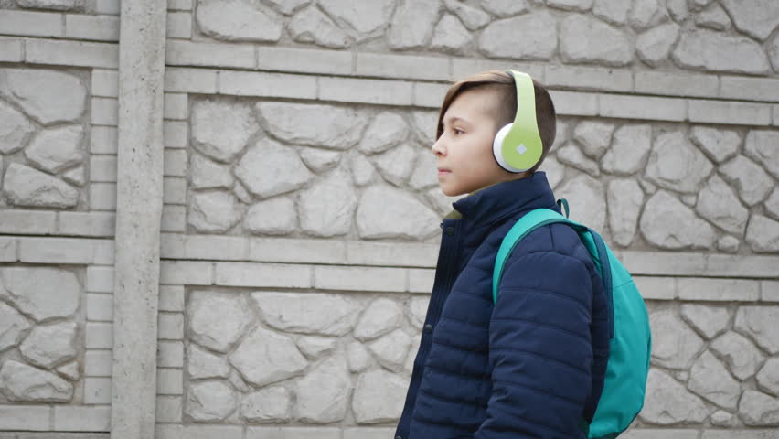 Handsome young man with a beautiful haircut in light green headphones and a green backpack walks next to a concrete fence