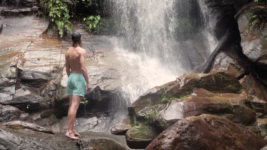 Young fit caucasian man in swimwear standing on rocks admiring a waterfall in Asia in slow motion camera tilting up