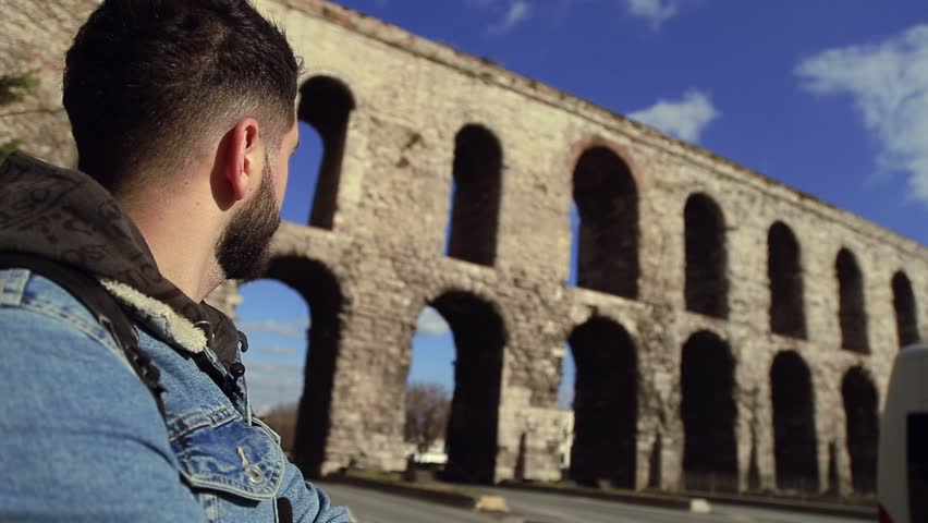 Close up view of young handsome bearded man in casual outfit looking at the ruins of old ancient fortress. Exploring history, architecture, travelling. Male portrait, few shots