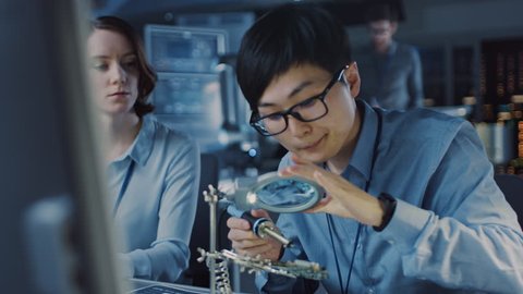 Japanese Development Engineer in Blue Shirt is Soldering a Circuit Board in a High Tech Research Laboratory with Modern Equipment. His Colleague Asks Him a Questio and Points on the Compter Screen.
