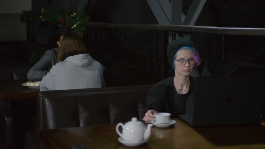 Tracking right of young hipster-woman sitting in bar at computer, typing on keyboard and drinking hot tea