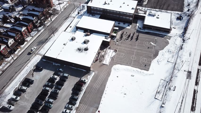 Aerial drone clip of an elementary school with children on the blacktop.  Shot on a sunny day in winter, snow on the ground.