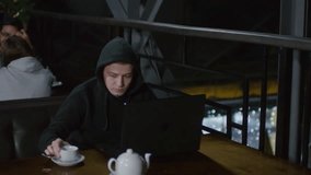 Sequence of videos with young Caucasian man in hoodie sitting at café table at laptop, coding and drinking tea