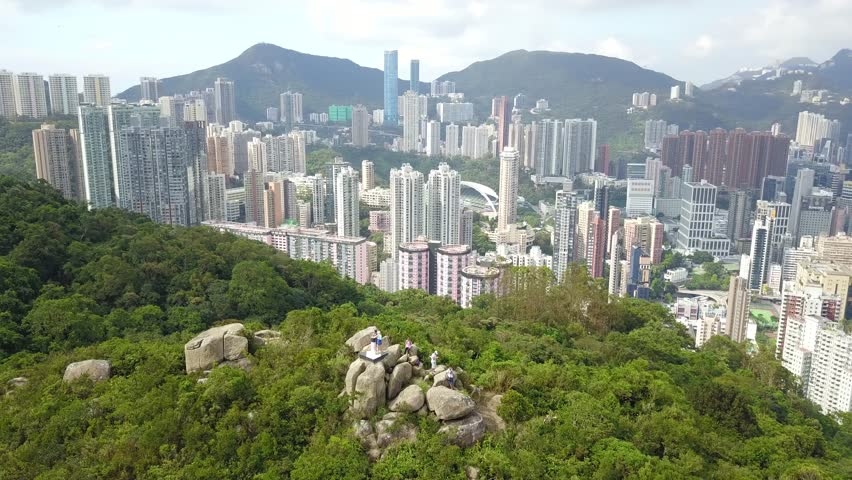 Aerial panorama of Hong Kong viewed from top of Braemar Hill Peak, with a city skyline of crowded skyscrapers by Victoria Harbour, residential towers on hillside & Kowloon downtown across the seaport