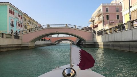 Venetian bridge on canals of picturesque Qanat Quartier icon of Doha, Qatar from a touristic boat with flag of Qatar. Venice at the Pearl, Persian Gulf, Middle East. Famous attraction at sunset light.