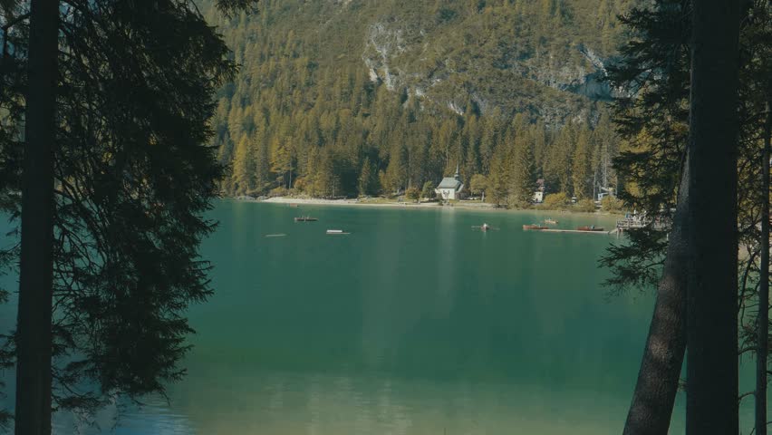 Looking through trees at the stunning Pragser Wildsee with forest covering a mountain hillside and a turquoise blue lake with boats and a house in the distance