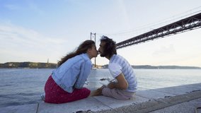 Happy man and woman kissing and taking off drone from hands while sitting on stone seafront on background of bridge