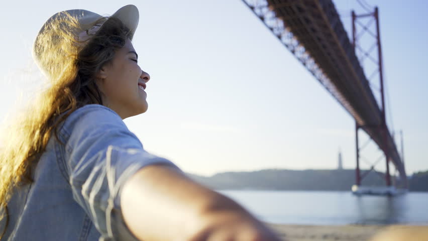 Point of view attractive happy lady in hat holding person hand on embankment near water under bridge in Lisbon