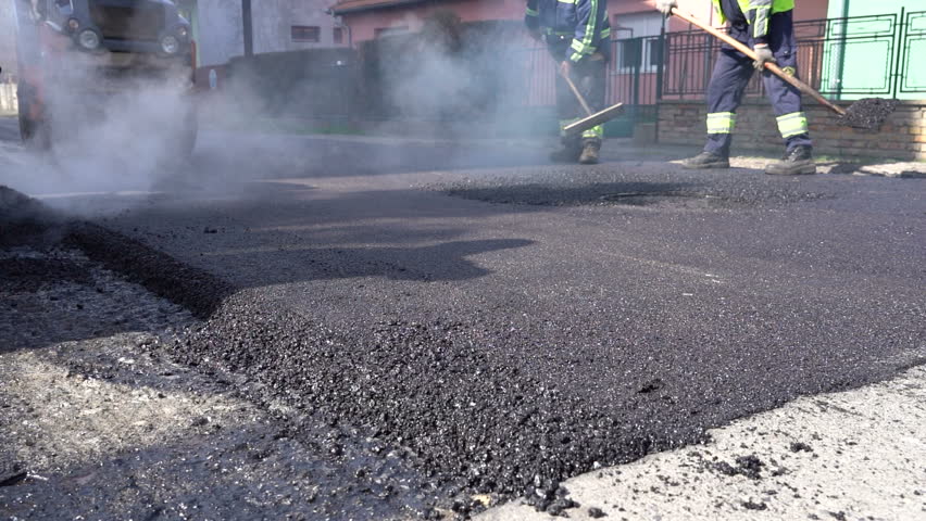 Constructions Workers Working on Road Construction - Slow Motion. Steam is Rising From The Hot Asphalt Surface. Worker Leveling Fresh Asphalt On A Road Construction Site. Royalty-Free Stock Footage #1026476243
