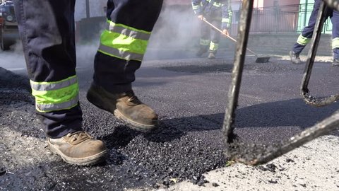 Constructions Workers Working on Road Construction - Slow Motion. Steam is Rising From The Hot Asphalt Surface. Worker Leveling Fresh Asphalt On A Road Construction Site.