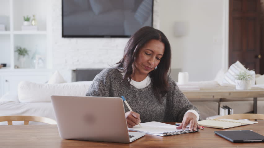 Middle aged woman sitting at a table at home making notes in a notepad and using laptop, front view Royalty-Free Stock Footage #1026478763