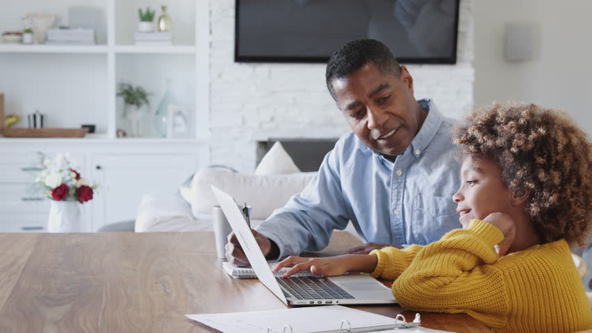 Pre-teen black girl sitting at a table working on a laptop computer with her home tutor, side view
