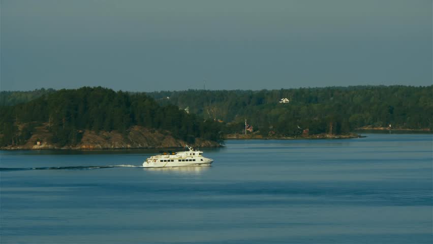 A yacht moves across calm waters of the Baltic among the islands of Stockholm Archipelago. Lush tree covered hills and islands in the background and clear sky on the horizon. Slow motion aerial view.