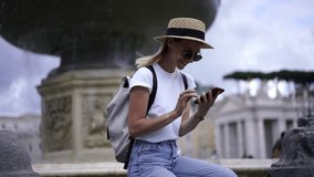 Happy hipster girl in sunglasses enjoying rest near fountain taking pictures of showplaces during vacations, positive female traveller using smartphone camera for photographing outdoors architecture