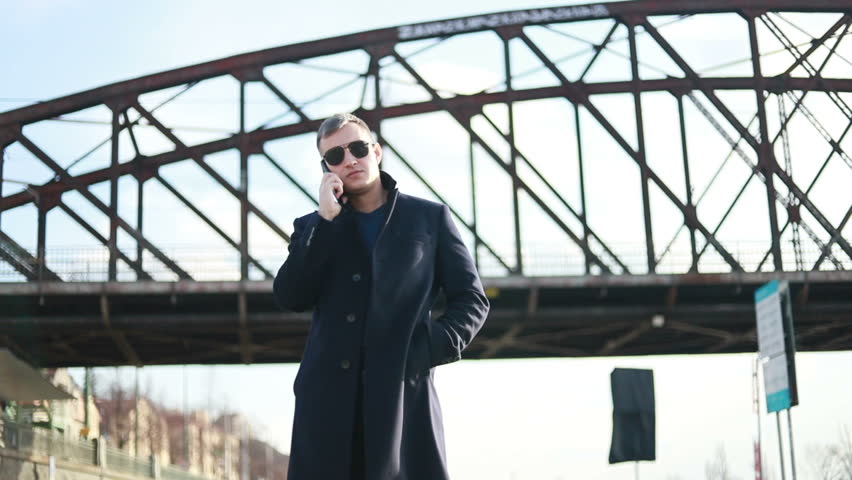 Young handsome businessman is talking on phone walking along street, back view of elegant man in suit is in business conversation, stand on background of cityscape. Concept: technology, business.