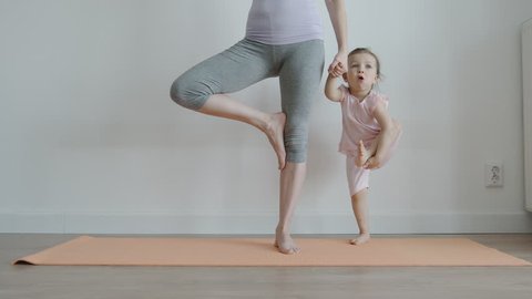YOGA: Slender mom with her pretty little daughter practising yoga on a mat at home - Front View