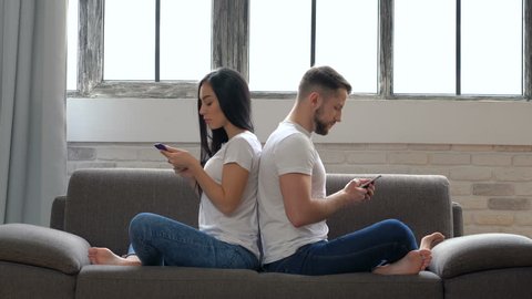 Modern couple sitting on the sofa back to back. No emotional, dont talking. Holding smartphones and looking at it. Couple problems. Communication problems.