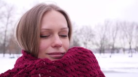 Portrait of a young blue-eyed pretty smiling woman in the winter city park who looking into the camera and wraps herself in a knitted red scarf in 4K slow motion close up video.