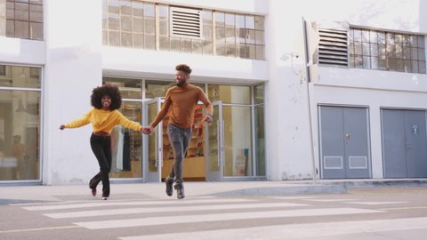 Fashionable young black couple running across a city street holding hands, full length