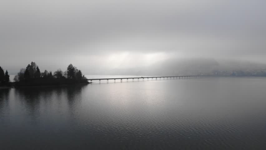 Dramatic low drone flight by low winter sunset light approaching the narrow bridge over the Einsiedeln lake in Switzerland