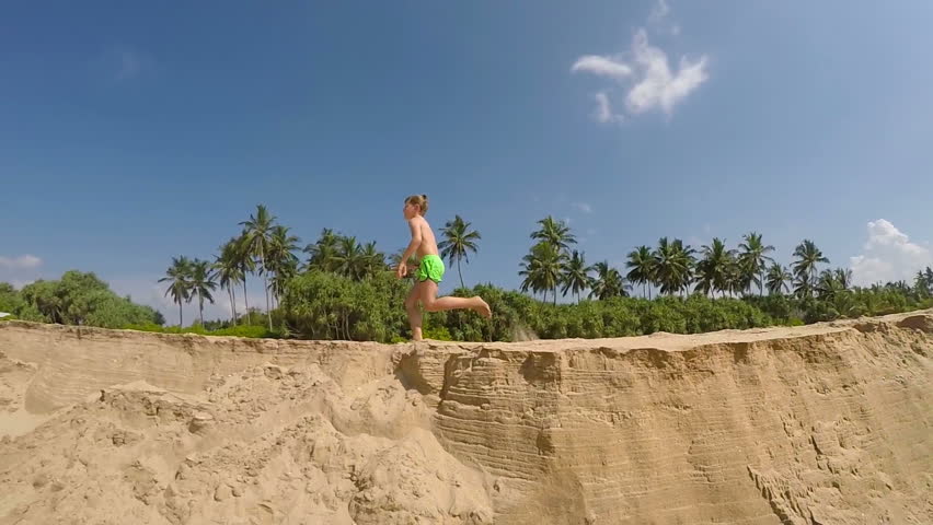 A boy runs on the sand beach with palms on the background slow motion